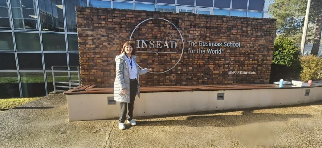 Karine wearing long puffy jacket and smiling, standing next to INSEAD Business School sign outdoors