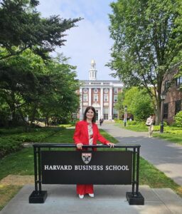 Karine wearing a bright red suit standing behind Harvard Business School sign outdoors on college campus