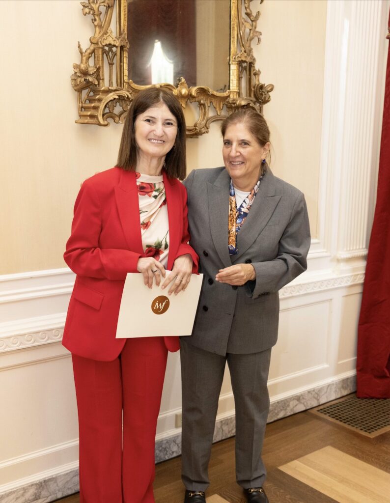 Karine stands with a woman in a grey suit who hands Karine a certificate in an ornate room