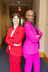 Karine wearing bright red suit standing back to back with a woman in a hot pink suit