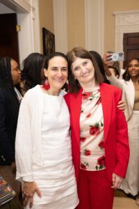 Karine wears bright red suit jacket, hugging a woman in a white dress with people mingling in background