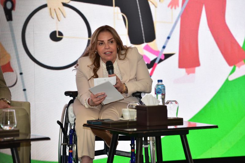 Asia speaks into a microphone on stage, wearing a suit and seated in wheelchair in front of colorful illustration depicting disabled figures