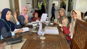 Five women with different disabilities sit around a table and smile at the camera, all holding up two-finger peace signs. Planning documents for their disability advocacy group project are spread out on the table.