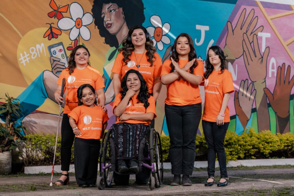 Six women with different disabilities pose together in matching orange WILD Guatemala shirts. They are all smiling and have their hair and make-up done expertly. Behind them in a colorful mural featuring a woman surrounded by flowers, butterflies, and hands reaching towards the sky.