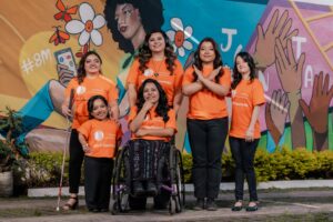 Six women with different disabilities pose together in matching orange WILD Guatemala shirts. They are all smiling and have their hair and make-up done expertly. Behind them in a colorful mural featuring a woman surrounded by flowers, butterflies, and hands reaching towards the sky.