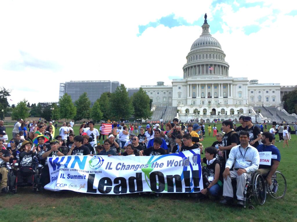 A large group of activists including exchange delegates from Pakistan gather in front of Capitol building in WAshington DC with large banner that says Independent Living Lead On