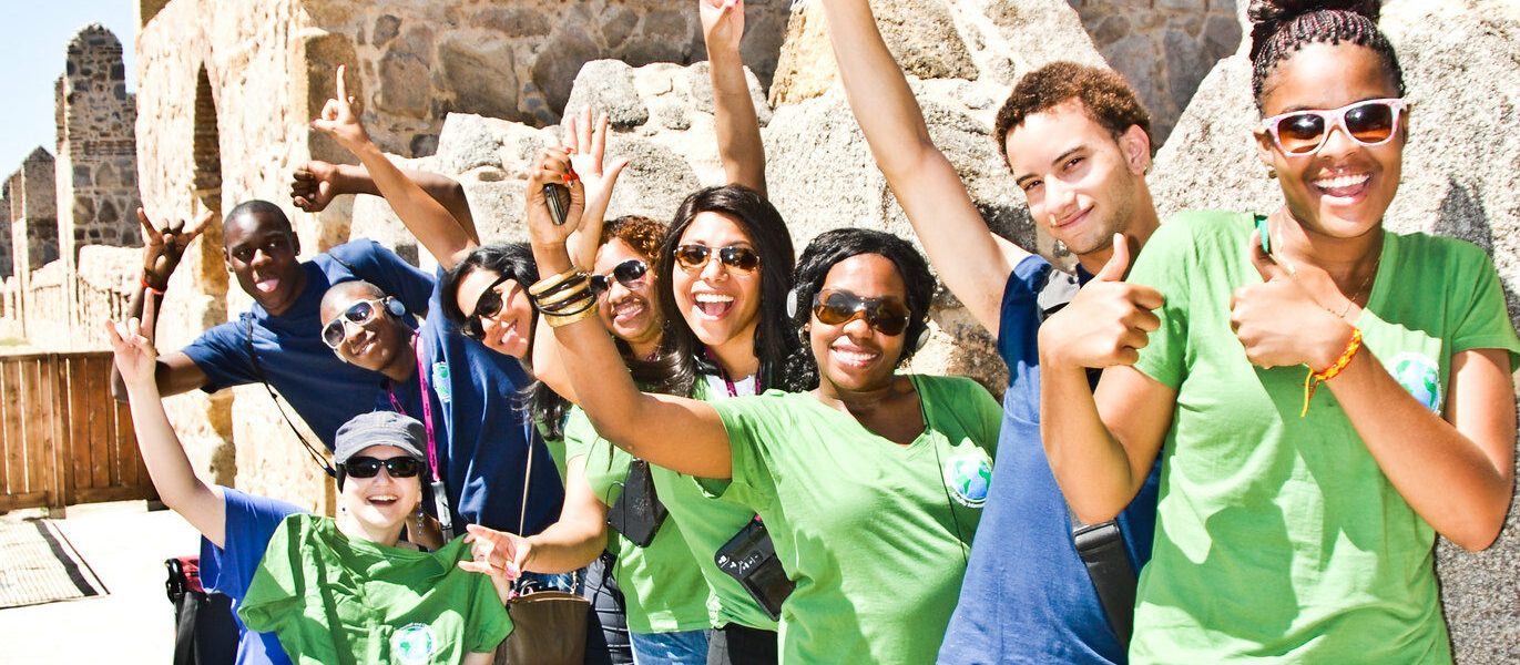A group of ten or so young people in front of a castle on a clear day smile and wave energetically at camera. Many are people of color and one person uses a power wheelchair while others stand.