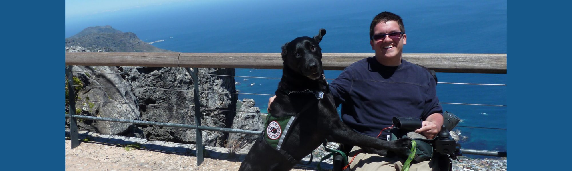 A young light skinned man in a powerchair, and his guide dog on Table Mountain in South Africa.